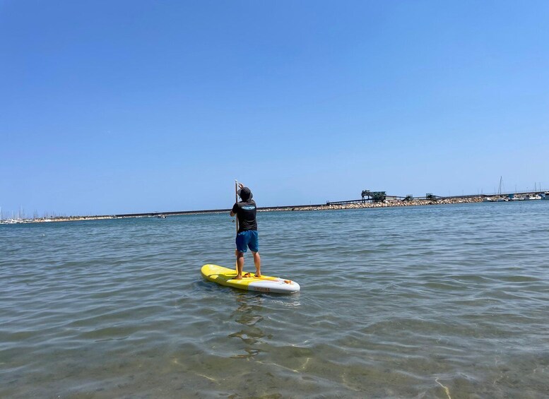 A person standing on a yellow paddleboard in calm water, with a clear sky and distant shoreline visible. 5