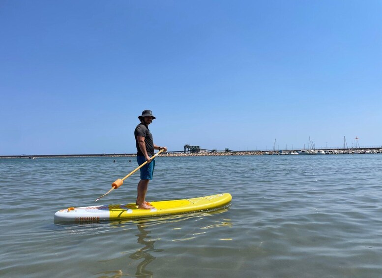A person paddleboarding on a calm body of water with a pier and boats in the background. 1