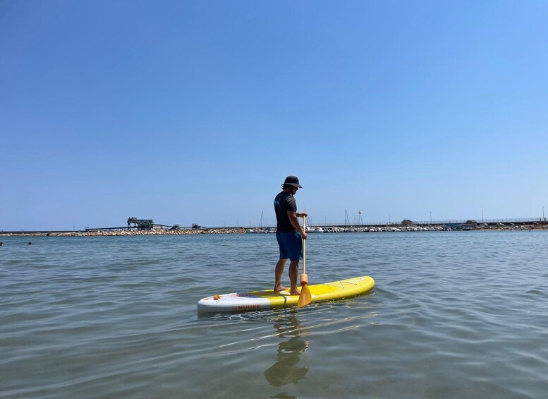 A person standing on a yellow paddleboard in calm water with a clear sky and a distant pier. 2