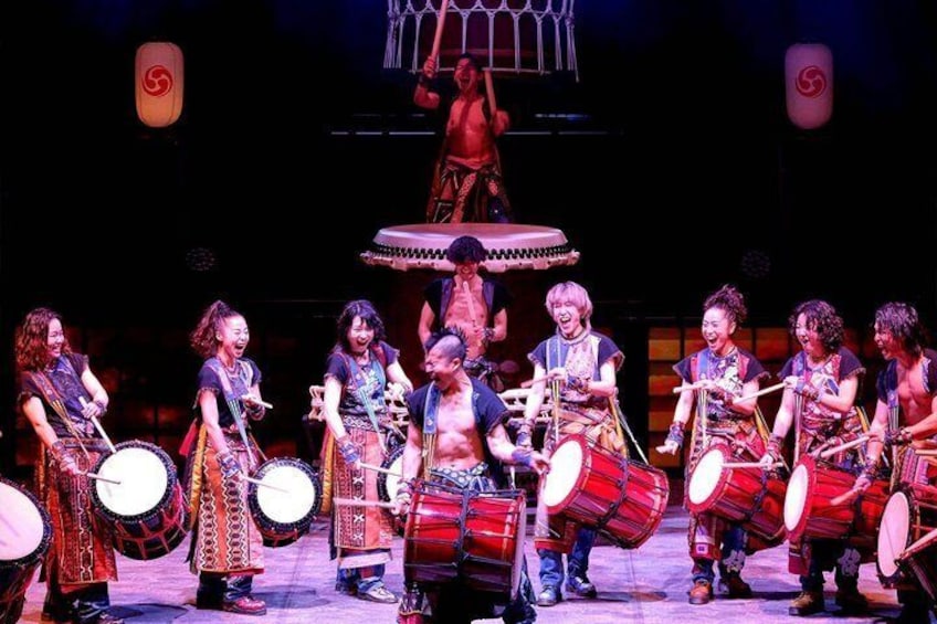 A group of performers on stage with drums and a central figure on a raised platform. 5