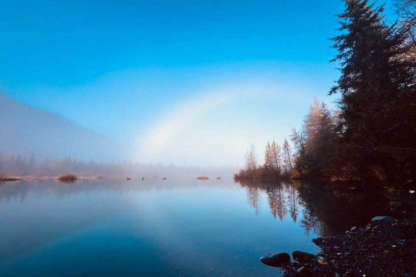 A calm lake with a rainbow, surrounded by trees and a mountain. 2