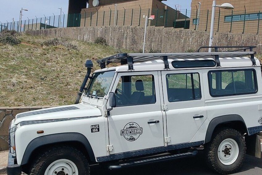 A white off-road vehicle with a roof rack parked on a paved area. 1