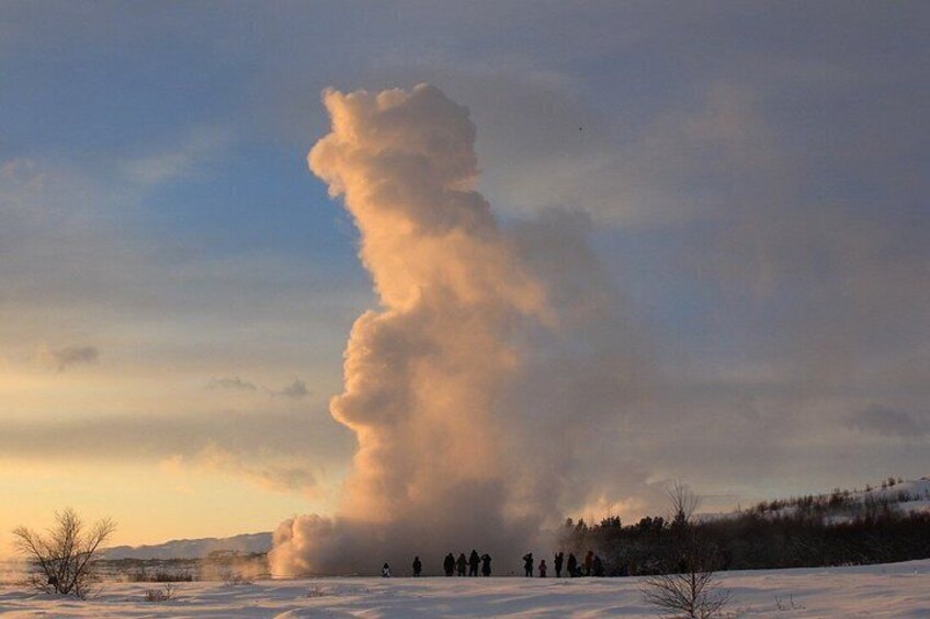 A snow-covered landscape with a large steam plume rising from the ground, surrounded by a group of people. 4