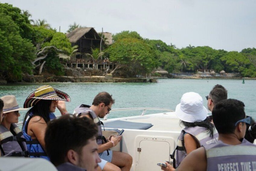 A group of people on a boat, some wearing life jackets, with a tropical landscape and a thatched-roof structure in the background. 1