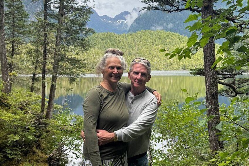 A couple embracing in a forested area with a lake and mountains in the background. 5
