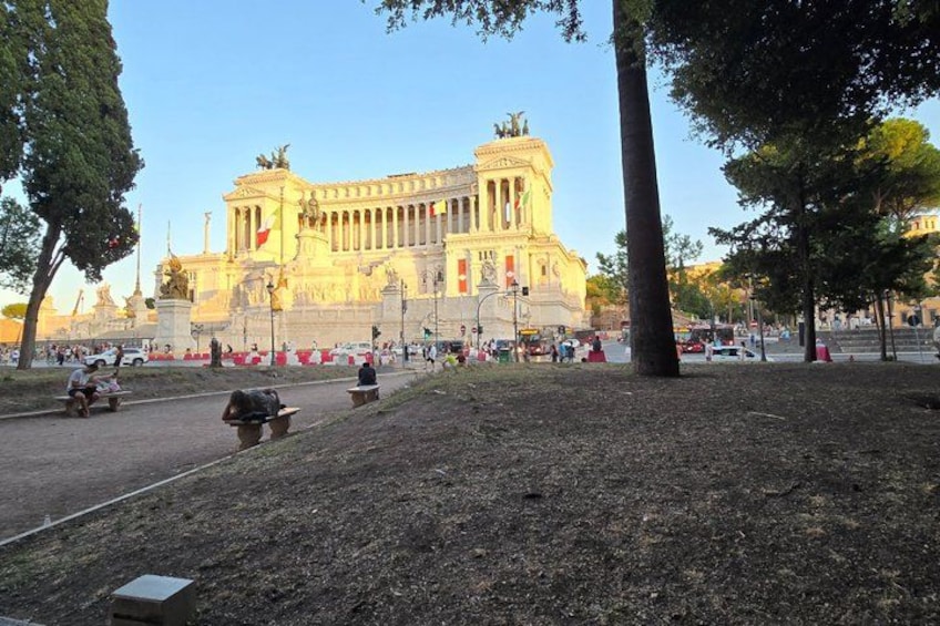 Ein großes, klassizistisches Gebäude mit Säulen und Statuen, umgeben von Bäumen und einer gepflasterten Fläche mit Menschen und Fahrzeugen. 2