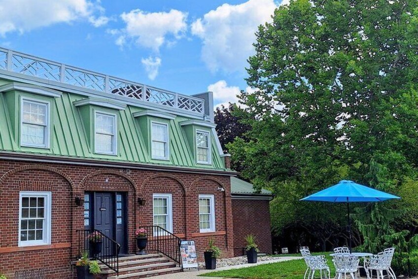 A brick building with a green roof, a black door, and white-framed windows. There is a blue umbrella and white chairs outside. 5