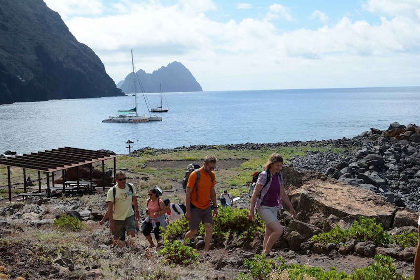 A group of people hiking near a rocky shoreline with boats in the background. 1