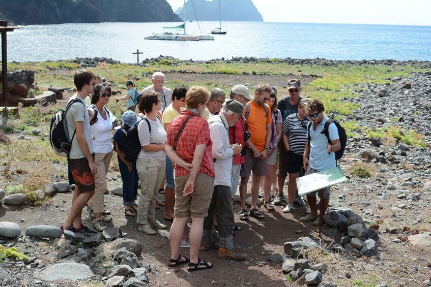 A group of people standing on a rocky beach with a sailboat in the background. 2