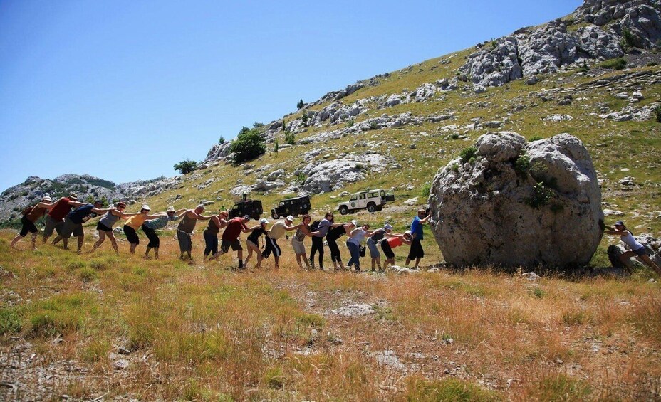 A group of people pulling a large rock across a grassy field with a mountainous backdrop. 5