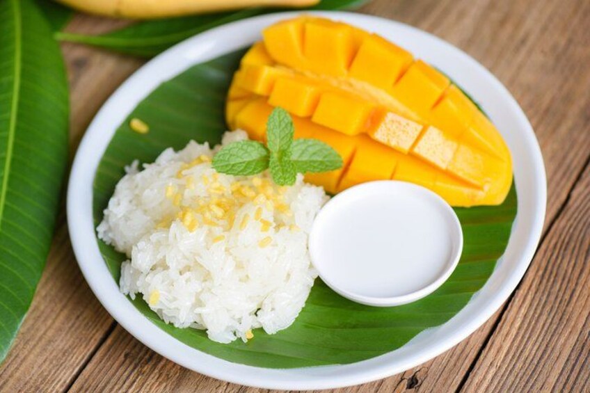 A plate of white rice with mango slices and a small bowl of sauce on a banana leaf. 4