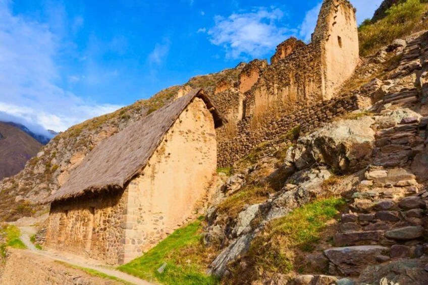 Ancient stone ruins with a thatched-roof structure, a steep rocky hillside, and a clear blue sky. 4