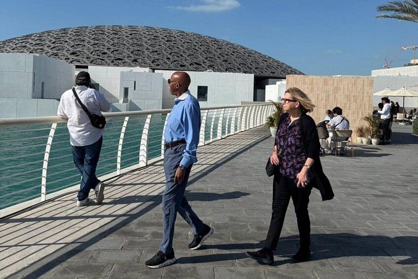 A waterfront promenade with people walking, a body of water, and a modern building with a unique roof design. 2