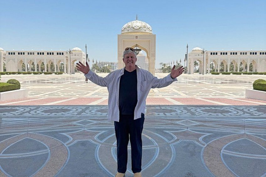 A person standing in a courtyard with geometric patterns on the ground, a large white building with arches in the background, and a clear blue sky. 5
