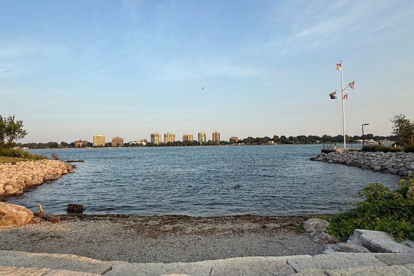A waterfront with a rocky shoreline, calm water, and a distant city skyline with buildings and flags. 3