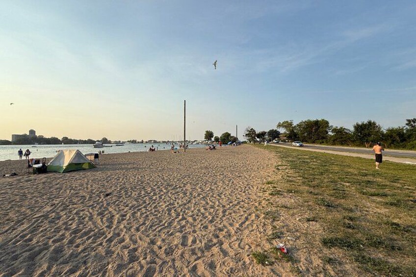 A sandy beach with a tent, a grassy area, a road, and a body of water in the background. 2