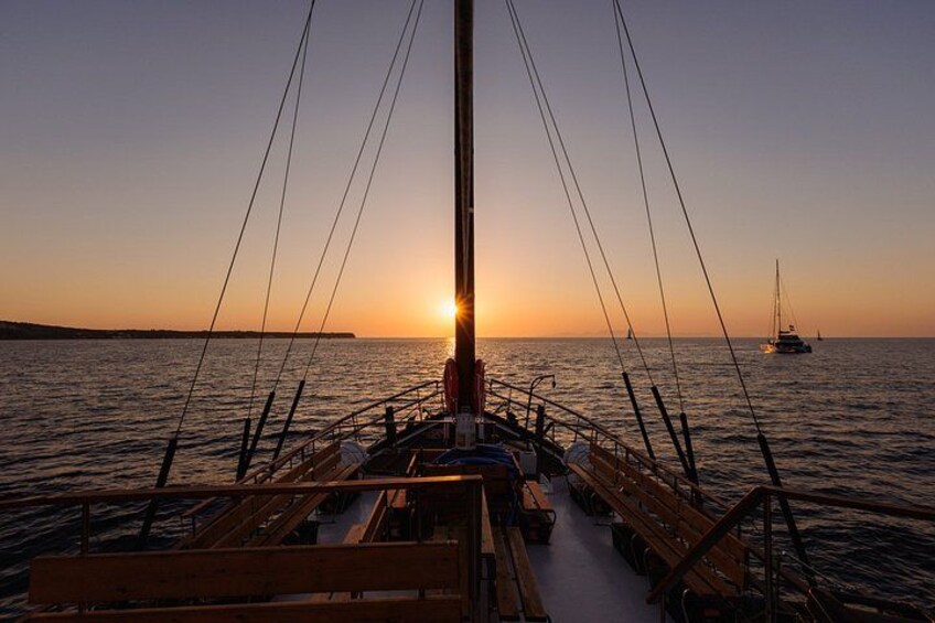 A boat at sunset with the sun setting on the horizon, a distant sailboat, and calm water. 1