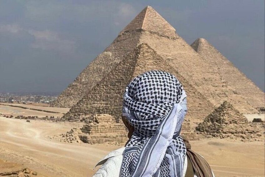 A person wearing a patterned headscarf and a striped garment, standing in front of the Great Pyramid of Giza. 3