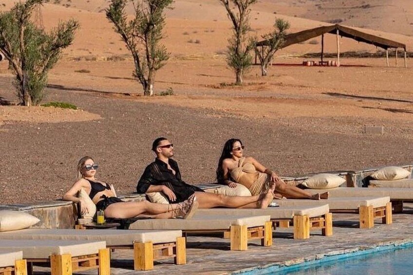 Three people relaxing on wooden lounge chairs by a pool in a desert setting. 3