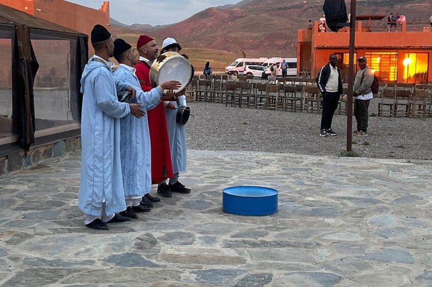 A group of people playing drums outdoors on a stone-paved area with mountains in the background. 3