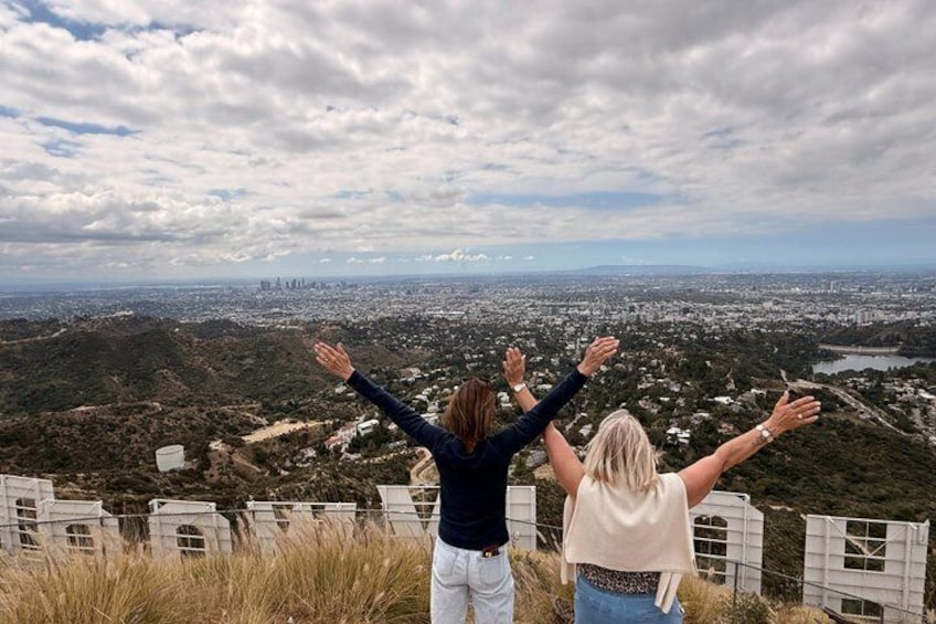Two people standing with arms raised, overlooking a cityscape with hills and a fence in the foreground. 4