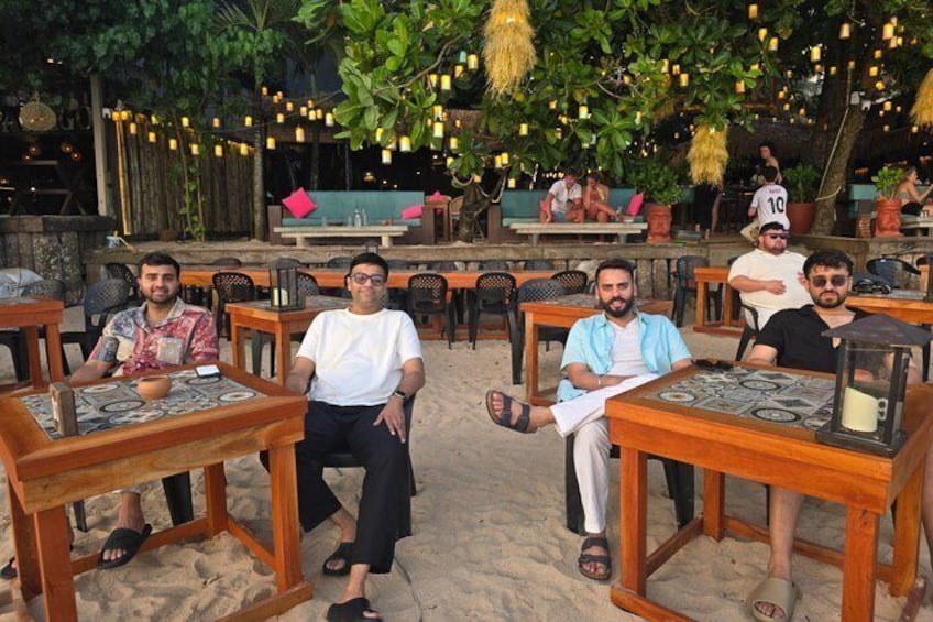 A group of people seated at wooden tables on a sandy beach with hanging lights and greenery in the background. 5