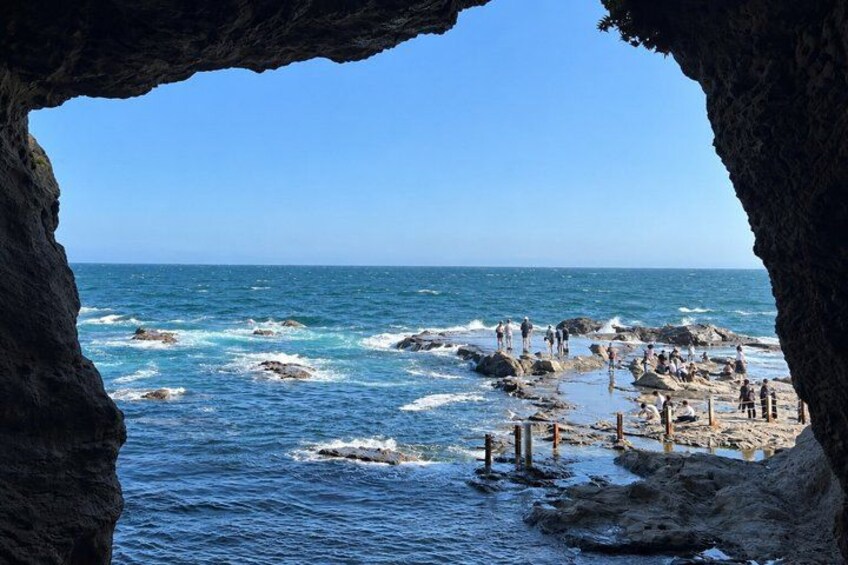 A rocky coastline with waves crashing against the shore, a clear blue sky, and people walking along the beach. 1