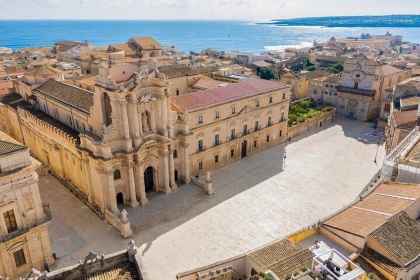 An aerial view of a coastal town with a prominent historical building, a large open plaza, and the sea in the background. 1
