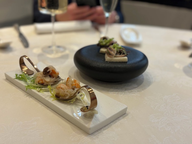 A plate of sushi with a decorative metal ring, a black stone bowl, and a glass of wine on a table. 5