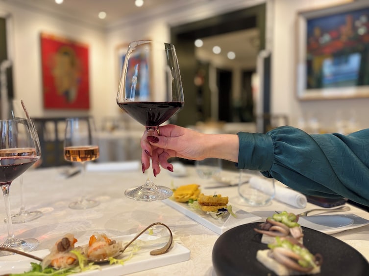 A person holding a wine glass with a red liquid, a dining table with various dishes, and a painting on the wall. 2
