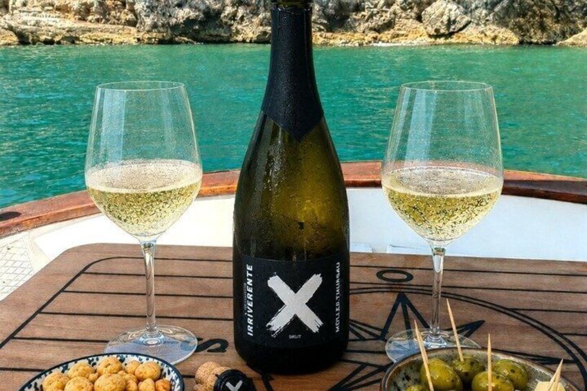 A bottle of white wine, two glasses, and a plate of olives on a boat with a rocky coastline in the background. 1