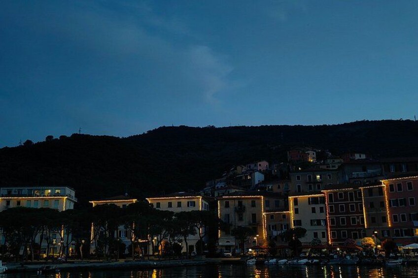 A coastal town at dusk with illuminated buildings and a hill in the background. 3