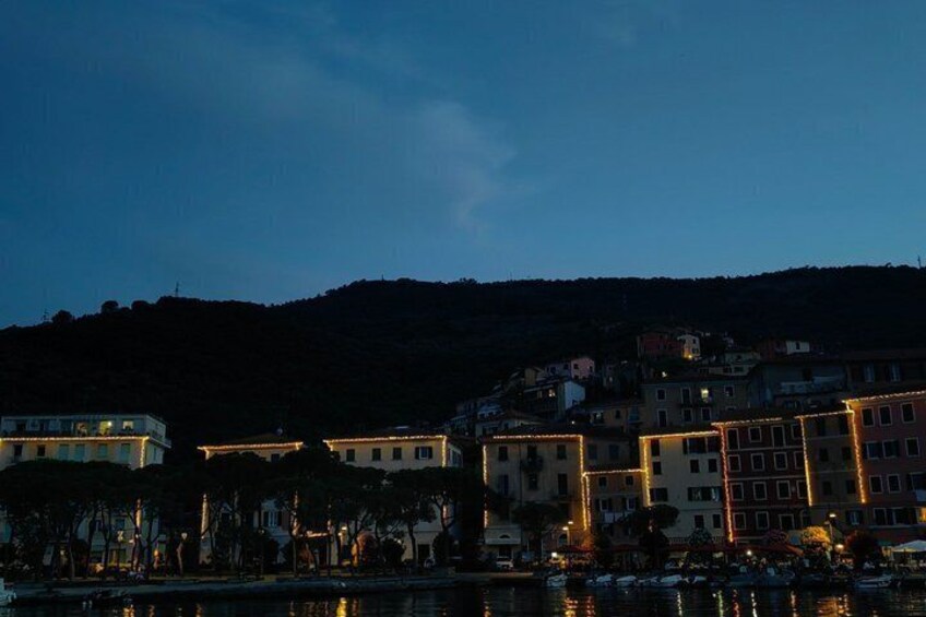 A coastal town at dusk with illuminated buildings and a hill in the background. 3