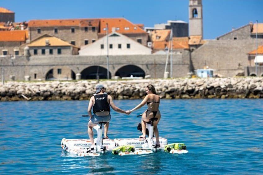Two people on a floating platform in a body of water, with buildings and a tower in the background. 4