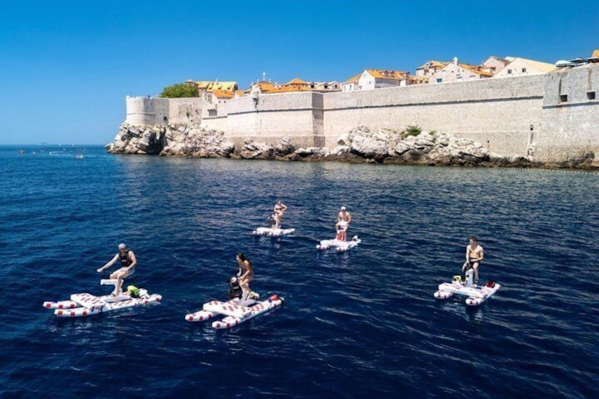 People on paddleboards in a body of water with a historic stone wall and buildings in the background. 1