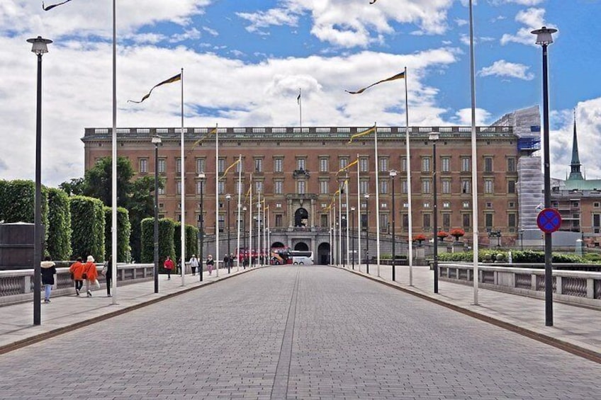 Un chemin pavé mène à un grand bâtiment orné de drapeaux, sous un ciel dégagé, et quelques piétons s'y promènent. 5