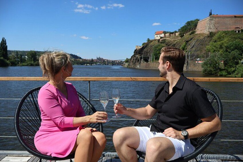 Two people sitting on a boat, holding wine glasses, with a river and a castle in the background. 4