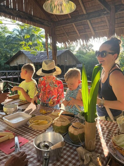 A family preparing food outdoors with a checkered tablecloth, a thatched roof, and a tropical setting. 2