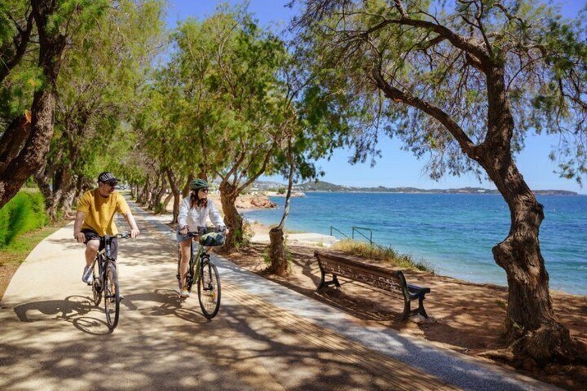 Two cyclists on a paved path with trees and a bench, overlooking a body of water. 3
