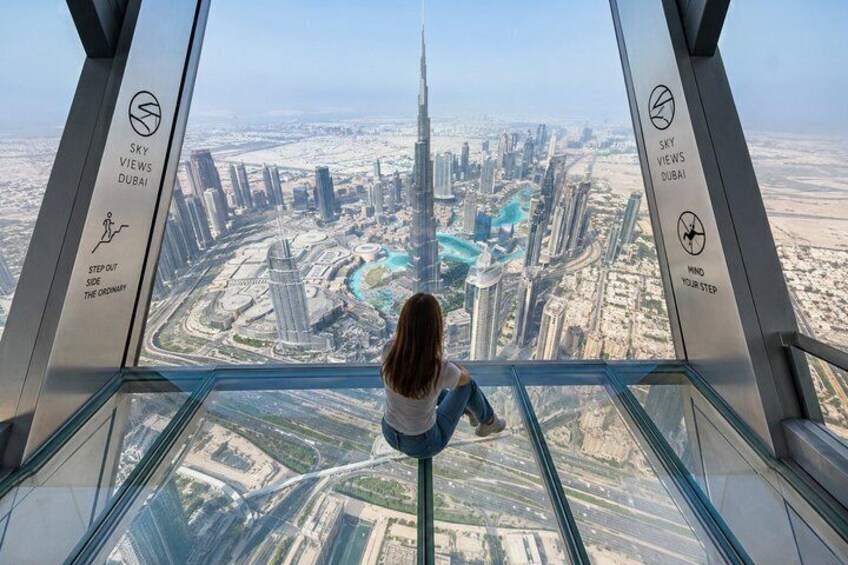 A person seated inside a glass observation deck with a view of a cityscape, including a prominent skyscraper. 1