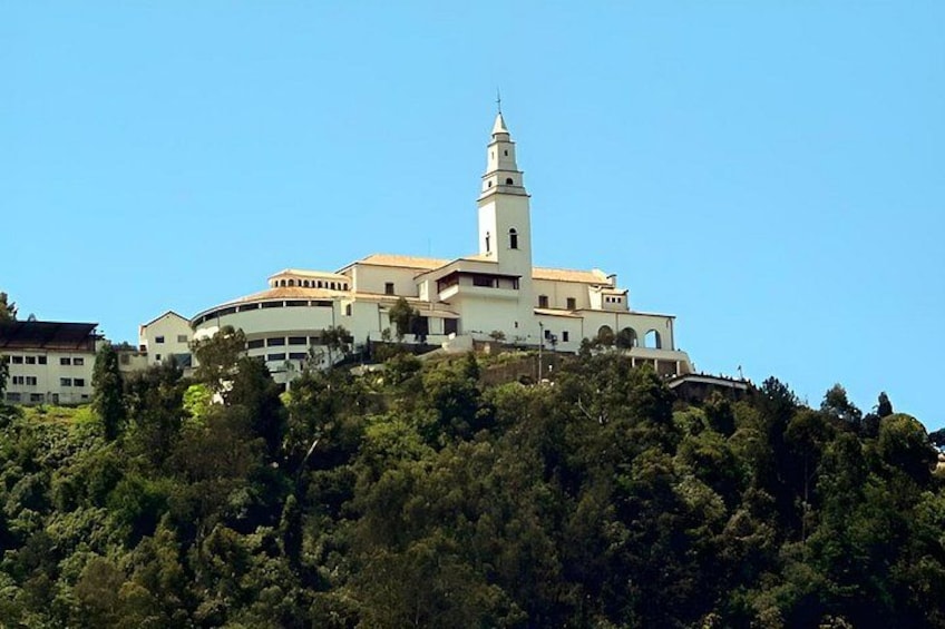 A white church with a bell tower on a hill, surrounded by dense green trees. 5