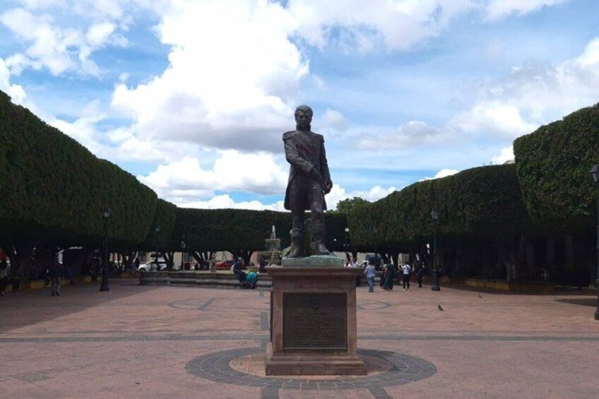 A statue in a square with a circular pattern on the ground, surrounded by trees and people. 4