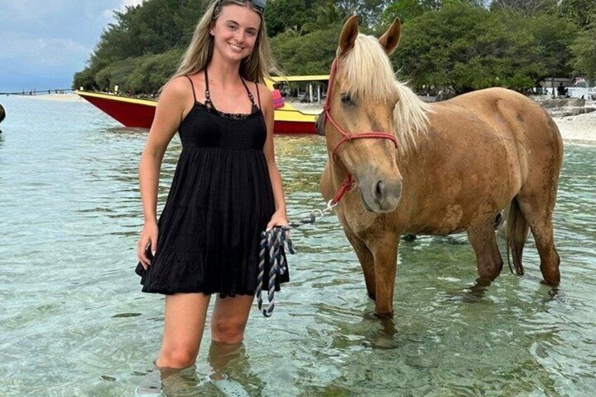 A person standing next to a horse in shallow water with boats and trees in the background. 4