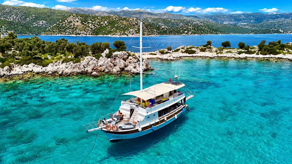 A boat named 'Leandris' anchored in clear turquoise waters with a rocky shoreline and a mountainous backdrop. 1