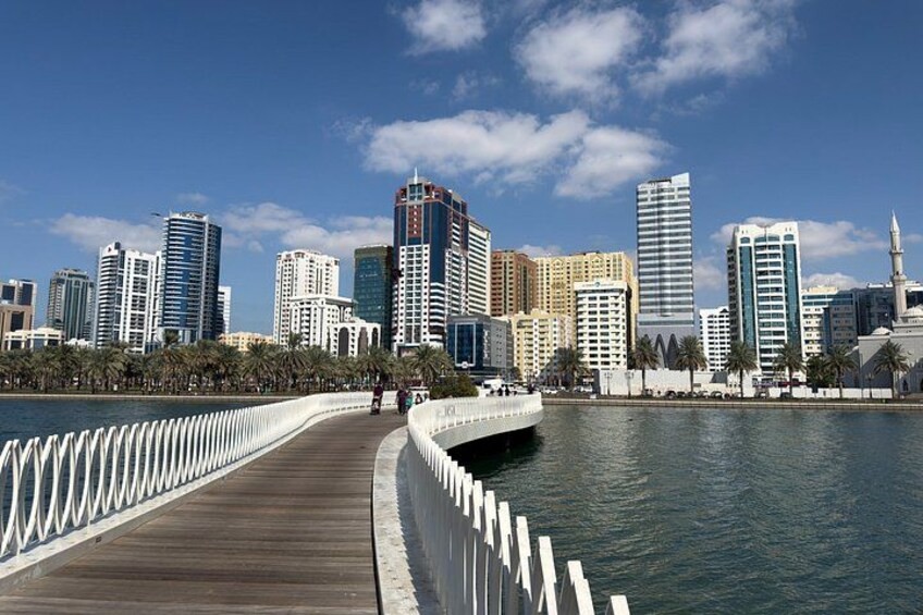 A waterfront promenade with a wooden walkway, palm trees, and a city skyline with modern high-rise buildings. 1