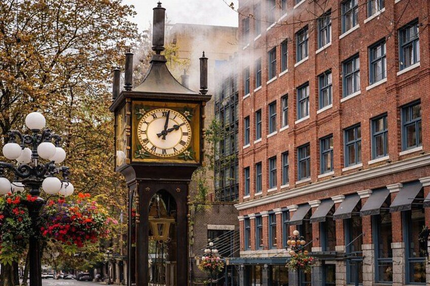 A clock tower with a clock face, surrounded by brick buildings and trees with autumn leaves. 3