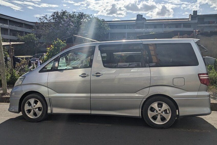 A silver minivan parked on a street with buildings and trees in the background. 3