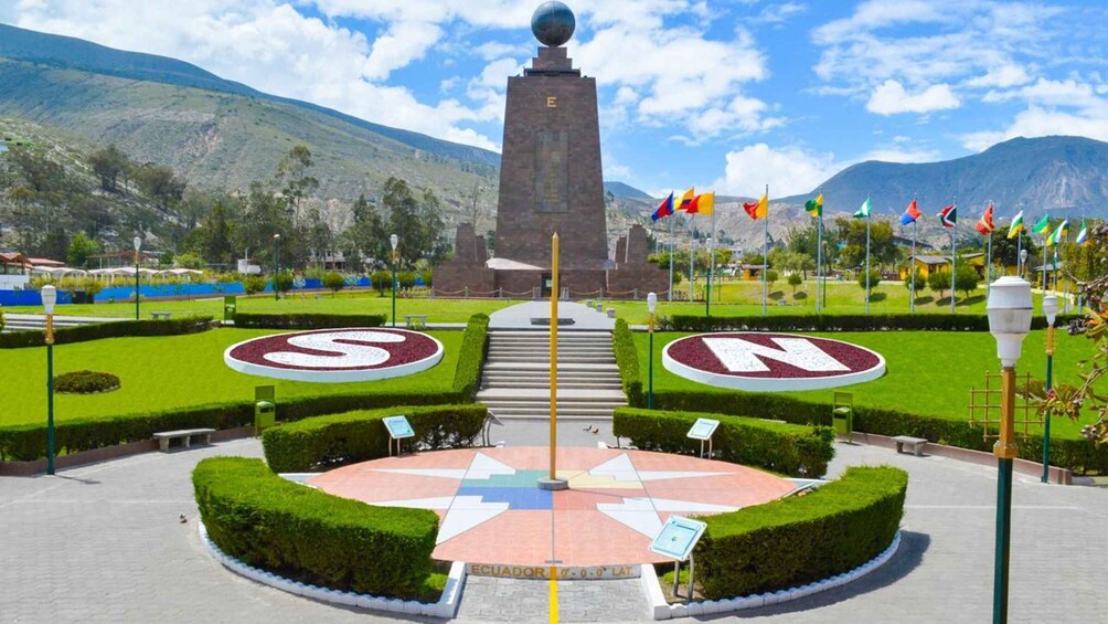 A monument with a globe on top, surrounded by a circular garden with compass-like designs, flags, and mountains in the background. 1