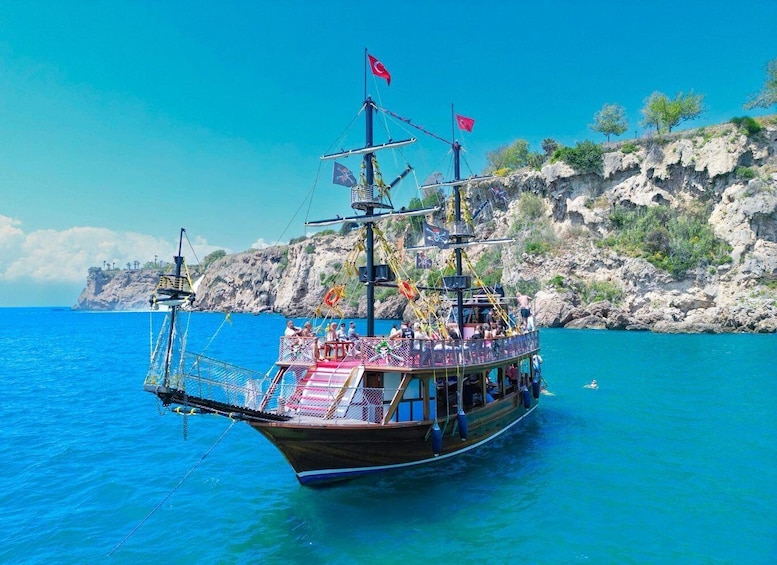 A traditional wooden boat with multiple flags, anchored in clear blue waters with rocky cliffs in the background. 3