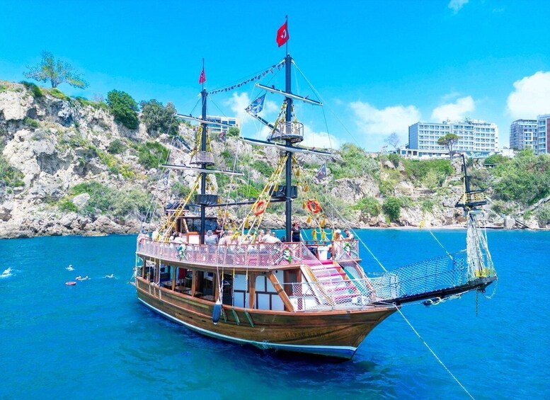 A wooden boat with a flag, anchored in a blue sea with rocky cliffs and buildings in the background. 5